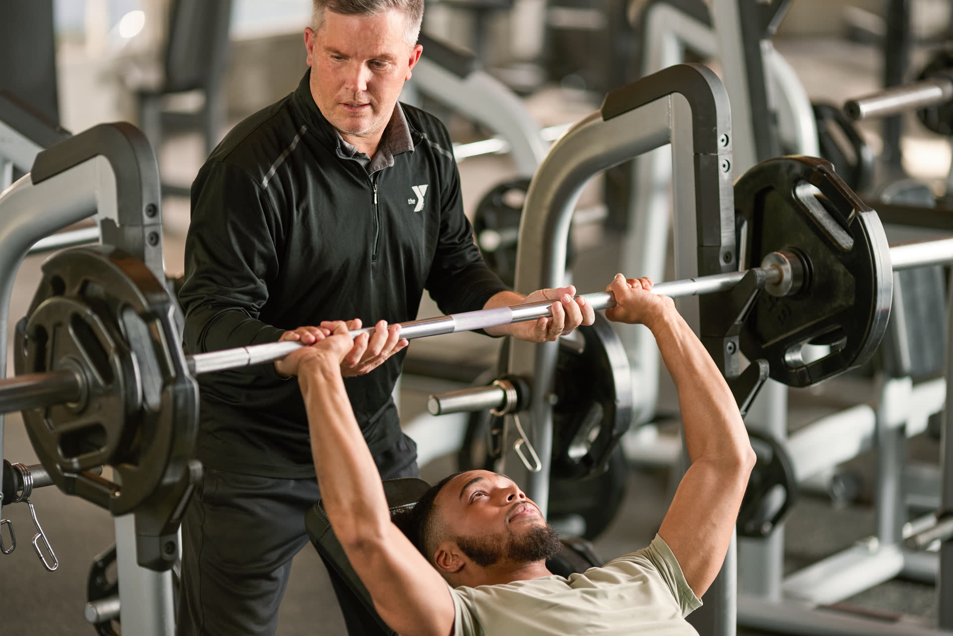 A YMCA staff member assisting a visitor lifting weights on a weight bench