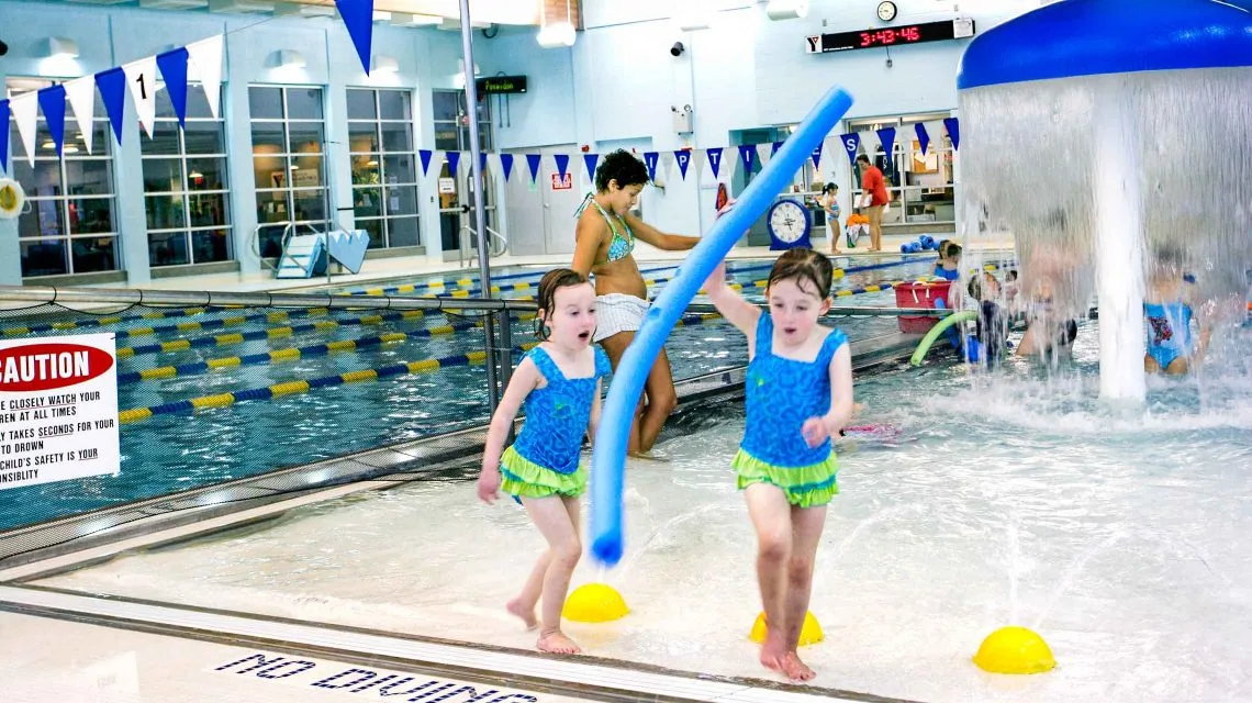 Children playing in a pool with a pool noodle and fountains in the background